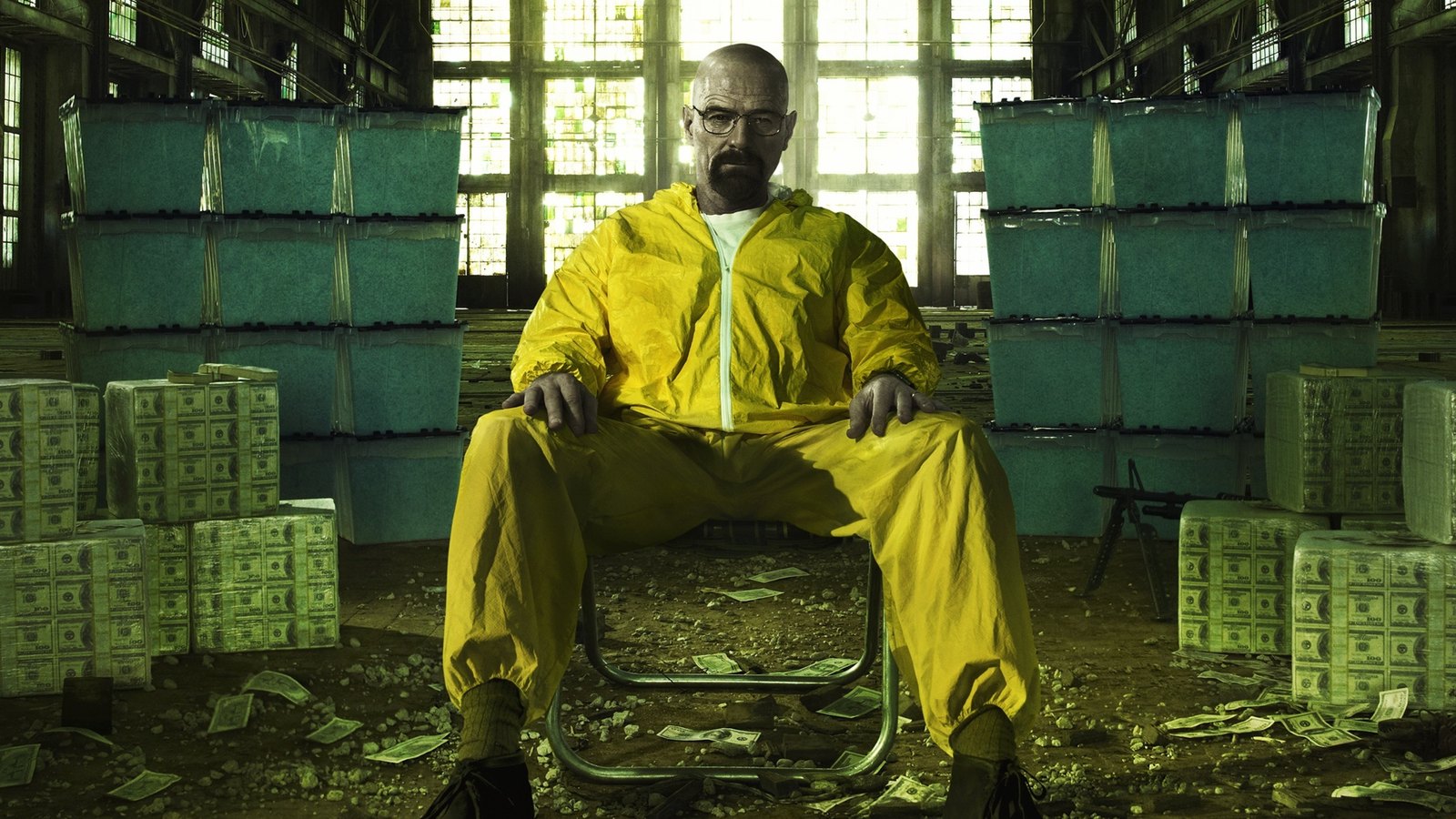 Walter White in a yellow hazmat suit seated amid stacked bins of product and bricks of cash in an abandoned industrial building.