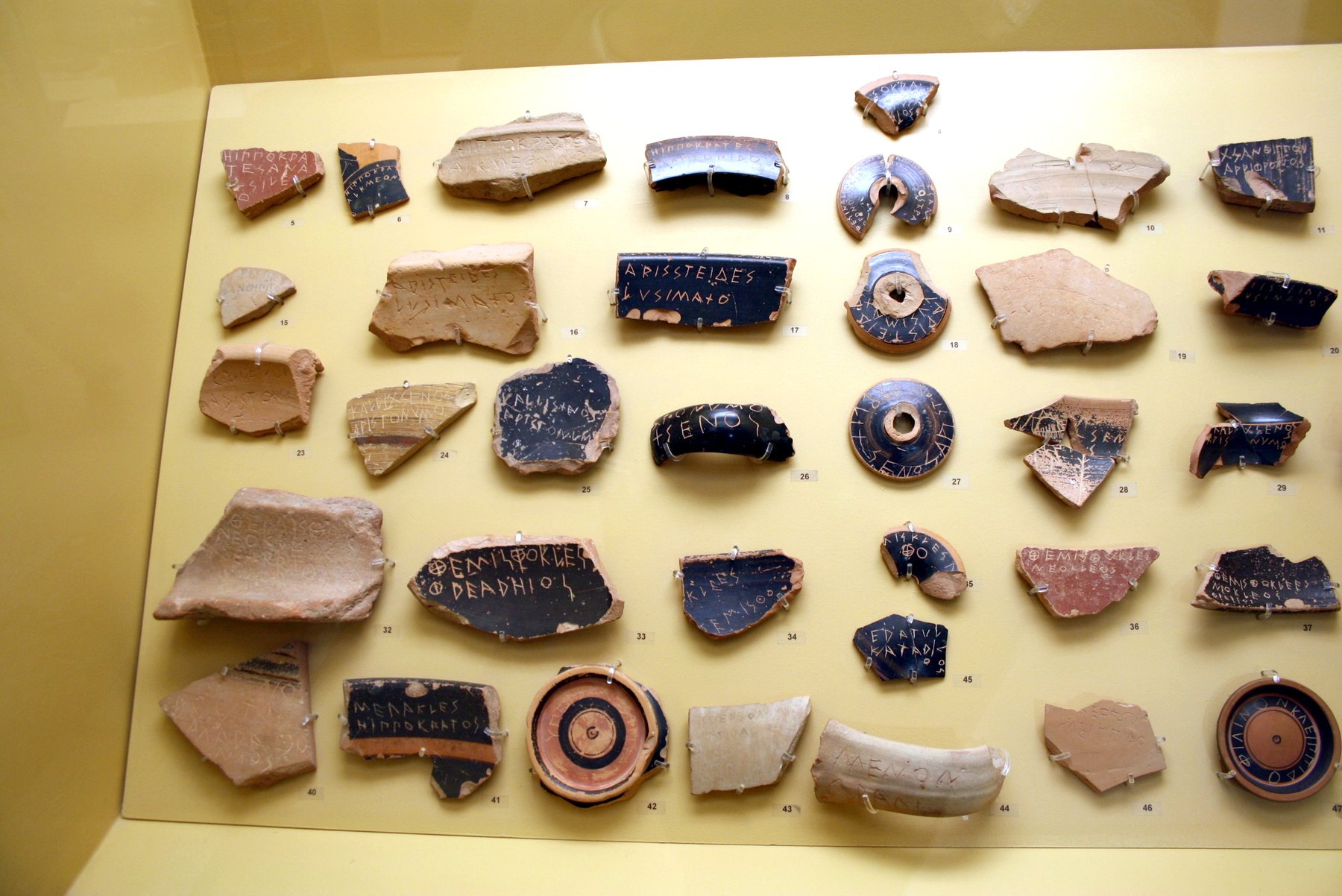 A museum case of ancient Greek pottery sherds used as ballots, each scratched with a politician's name in Greek capitals.